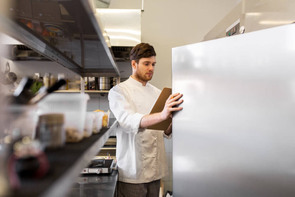 Male chef cook with clipboard doing inventory in restaurant kitchen fridge